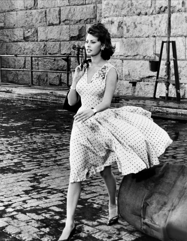 Sophia Loren wearing a 1950s polka dot summer dress, walking confidently on a cobblestone street — a timeless retro fashion icon and symbol of feminine elegance.

